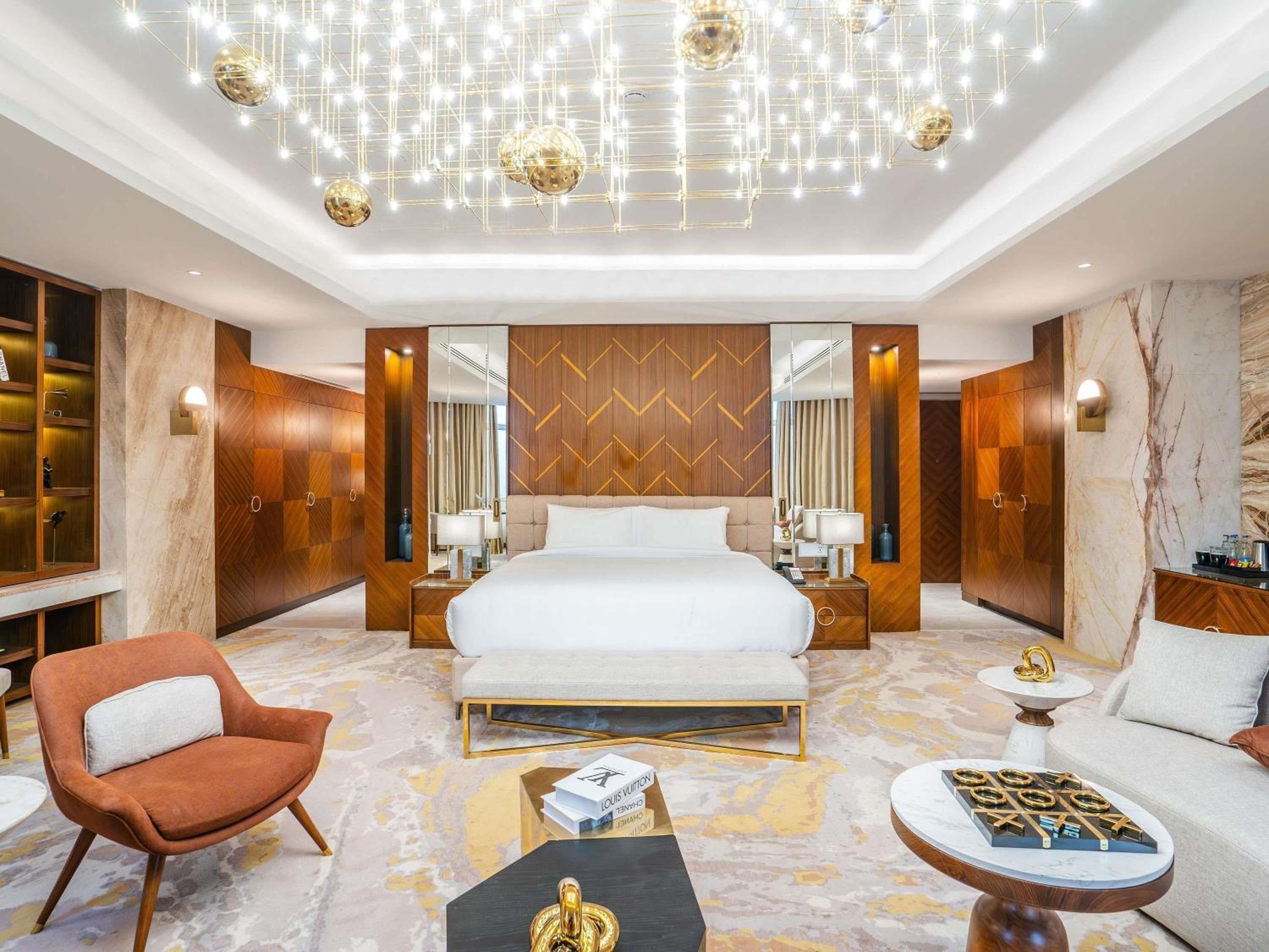 A modern hotel room with a large bed, a seating area with a sofa and armchair, a coffee table, and a chandelier.