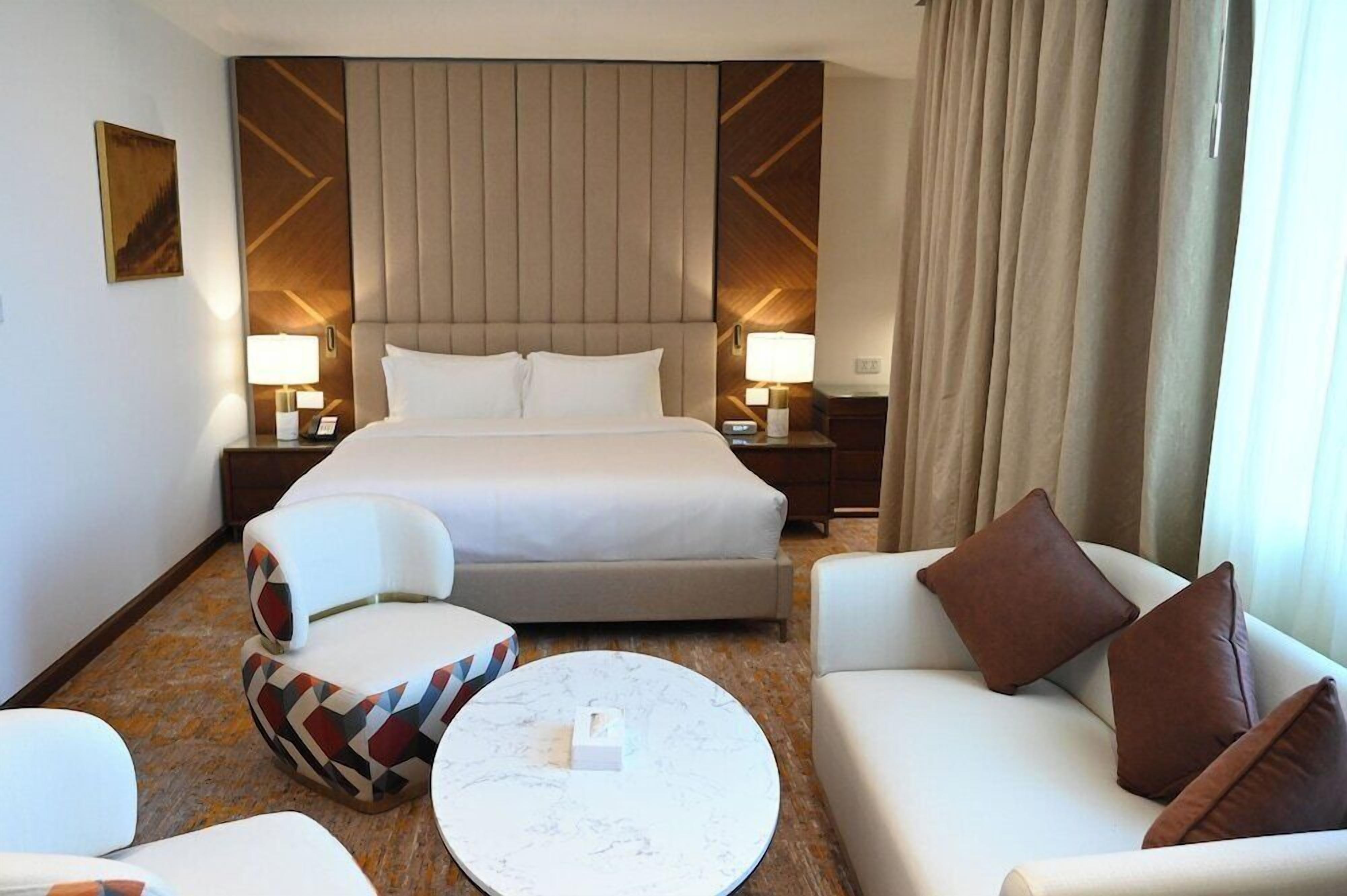 A hotel room with a large bed, two bedside tables with lamps, a sofa, a chair, and a coffee table.