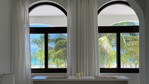 A room with a view of palm trees and the sea through arched windows, a white desk, and a lamp.