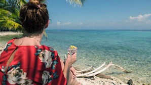 A person holding a drink by a beach with clear water and a palm tree.