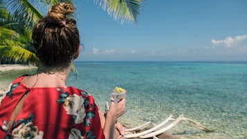 Iemand houdt een drankje vast aan een strand met helder water en een palmboom.