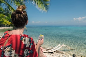 A person holding a drink by a beach with clear water and a palm tree. - The Hacienda - Studio West (Utila)