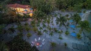 A beachfront property with a large house, a fire pit surrounded by lounge chairs, and a sandy beach with palm trees.