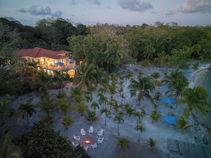 A beachfront property with a large house, a fire pit surrounded by lounge chairs, and a sandy beach with palm trees. - The Hacienda - Studio West (Utila)
