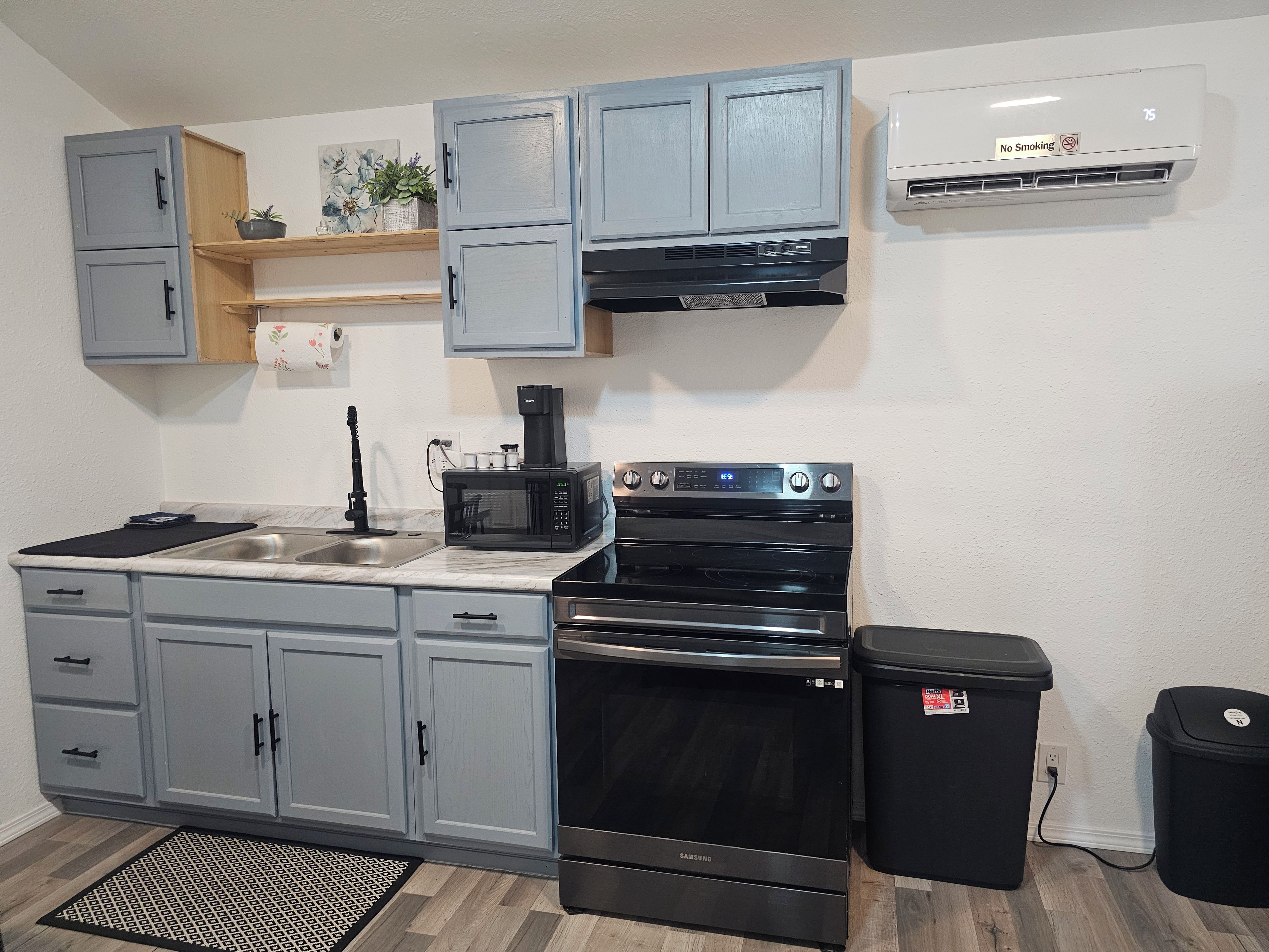 A compact kitchen with grey cabinets, a black microwave, a stainless steel sink, a black oven, and a trash bin.