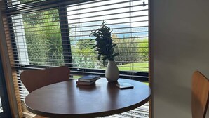 Mesa redonda con un jarrón y libros, rodeada de sillas, en una habitación con ventanas grandes y vistas a la vegetación.