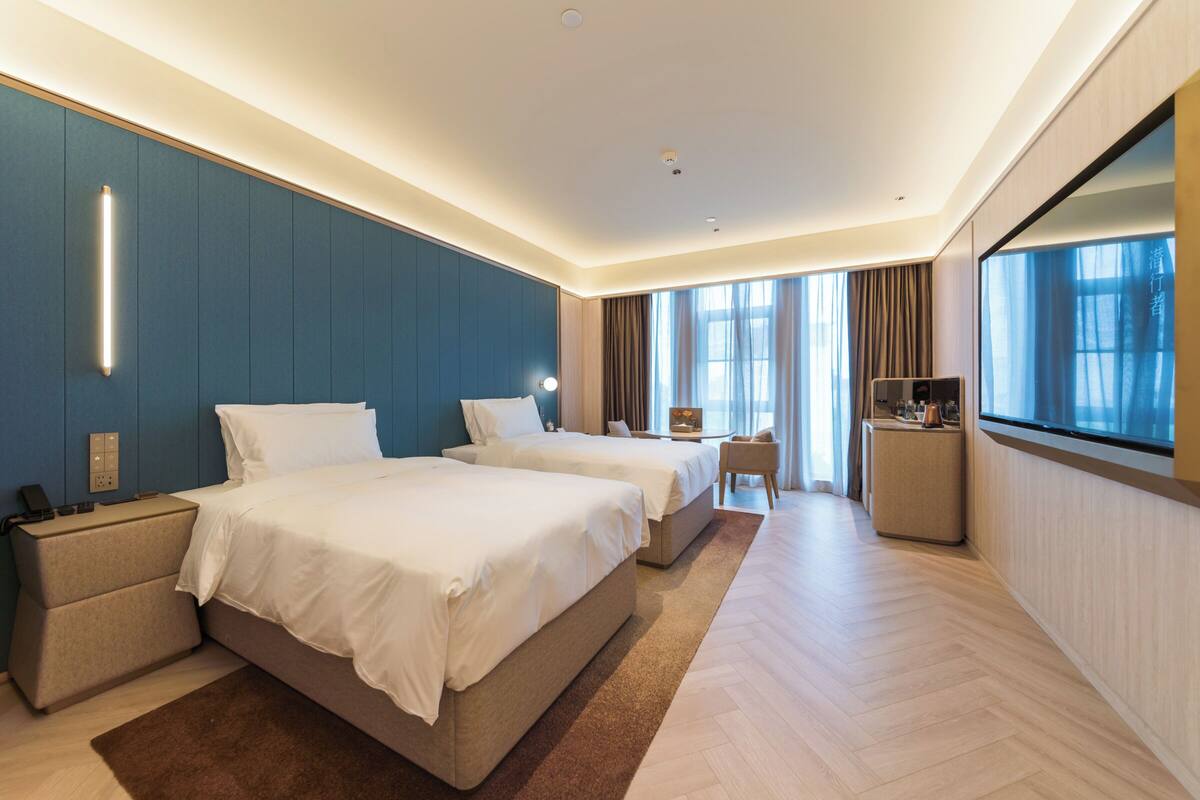 A hotel room with two beds, a desk, a chair, a TV, and a large window.