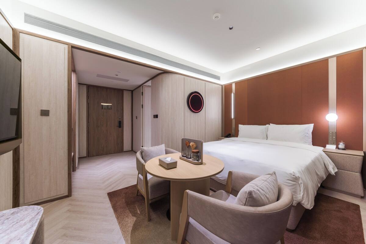 A hotel room with a bed, a desk, a chair, a TV, and a wall-mounted circular light.