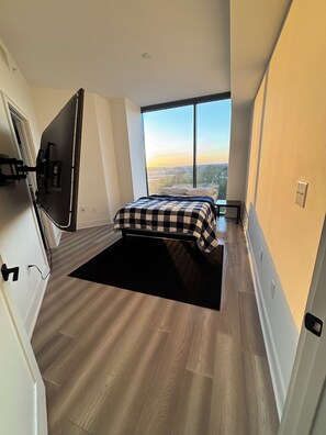 Ein Schlafzimmer mit großem Fenster, einem Bett mit kariertem Überwurf, einem schwarzen Teppich und einem an der Wand befestigten Fernseher.