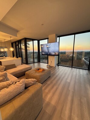 Ein modernes Wohnzimmer mit großem Fernseher, bequemen Sitzgelegenheiten und Blick auf die Stadt durch raumhohe Fenster.