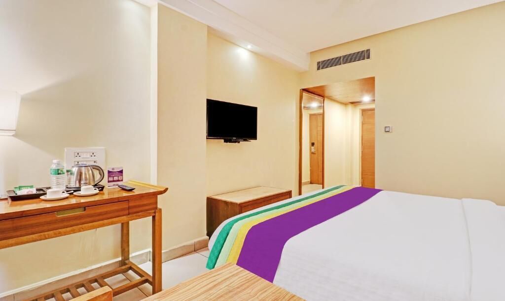 Quarto de hotel com cama, escrivaninha com chaleira, televisão de tela plana e porta de madeira.