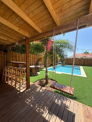 Eine Holzterrasse mit Pool, einer Schaukel und einem überdachten Sitzbereich.