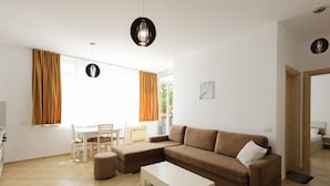 A modern living room with a sofa, ottoman, and coffee table, a dining area with chairs, and a large window with curtains. - Lura Apartment 05 in Plazhi San Pietro (Ishëm)