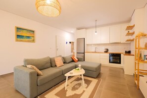 A modern living room with a sofa, a small coffee table, and a kitchen area with white cabinets and a wooden shelf. - Sanpietro Vacation Apartment 052 (Ishëm)