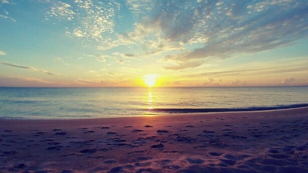 Ein Strand mit Fußabdrücken, die zum Wasser führen, ein Sonnenuntergang im Hintergrund und ein klarer Himmel.