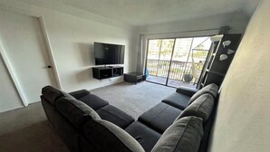 Ein Wohnzimmer mit einem Ecksofa, einem Flachbildfernseher und einer Glastür, die auf einen Balkon führt.