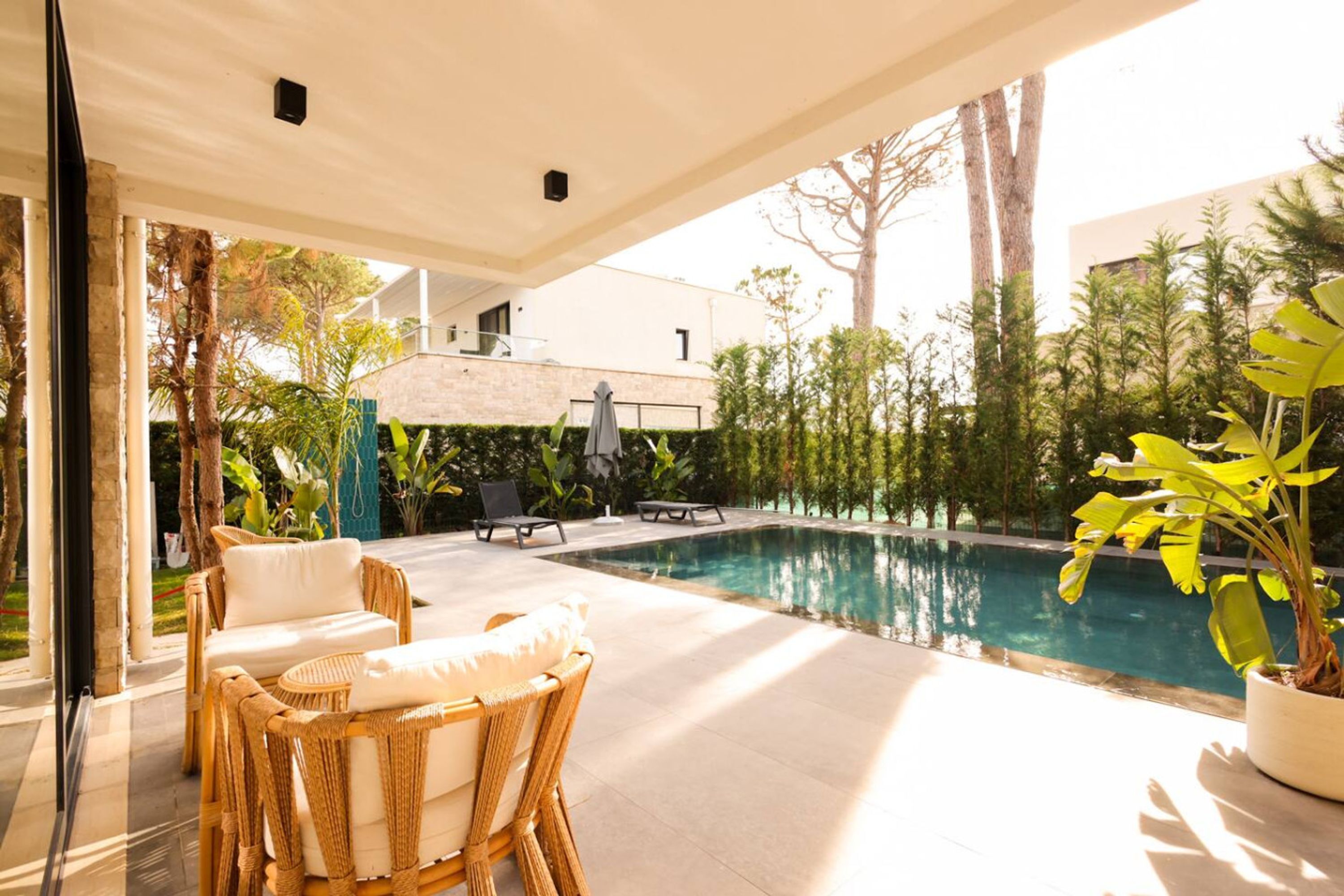 An outdoor pool area with lounge chairs, a covered seating area, and a view of a building and trees.