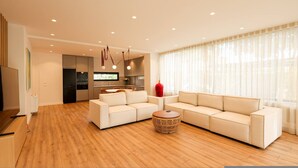Una sala moderna con un conjunto de sofás, una mesa de centro redonda de mimbre y una cocina al fondo.