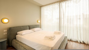 Un dormitorio con cama, lámparas de noche y una ventana con cortinas translúcidas.