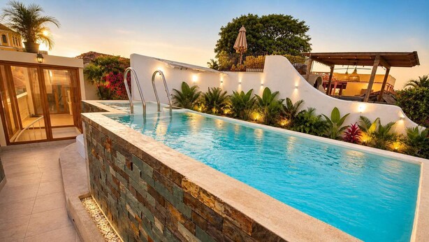 Une piscine avec un mur en pierre, une entrée avec une porte en verre et un espace abrité agrémenté de plantes.