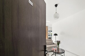 Ein modernes Interieur mit einer dunkelholznen Tür, einem Sofa, einem kleinen runden Tisch mit Getränken, einer Vase mit Blumen und einer hängenden Leuchte.