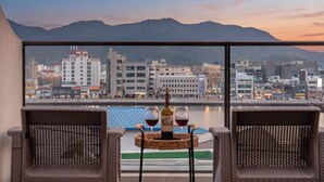 Quarto com vista para um horizonte urbano e montanhas, duas cadeiras de vime, uma mesa redonda pequena com uma garrafa e copos.