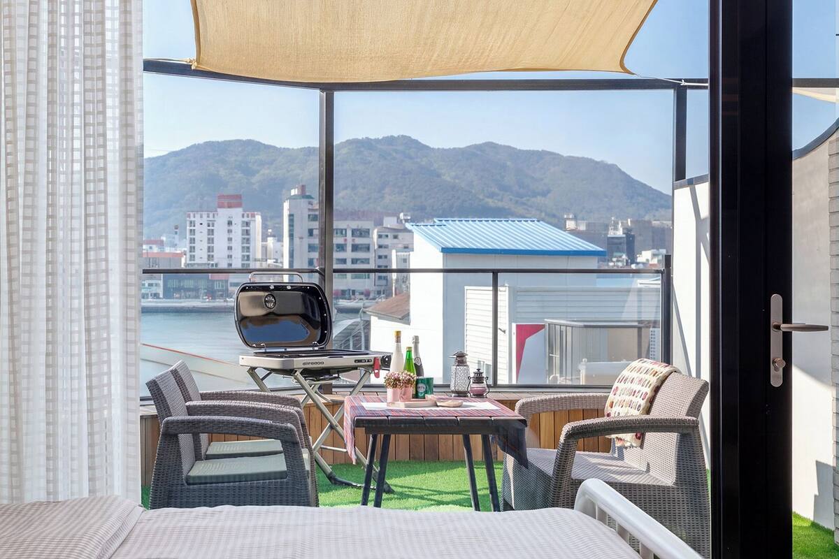 Un balcón con mobiliario de mimbre, una parrilla y vistas a un paisaje urbano y montañas.