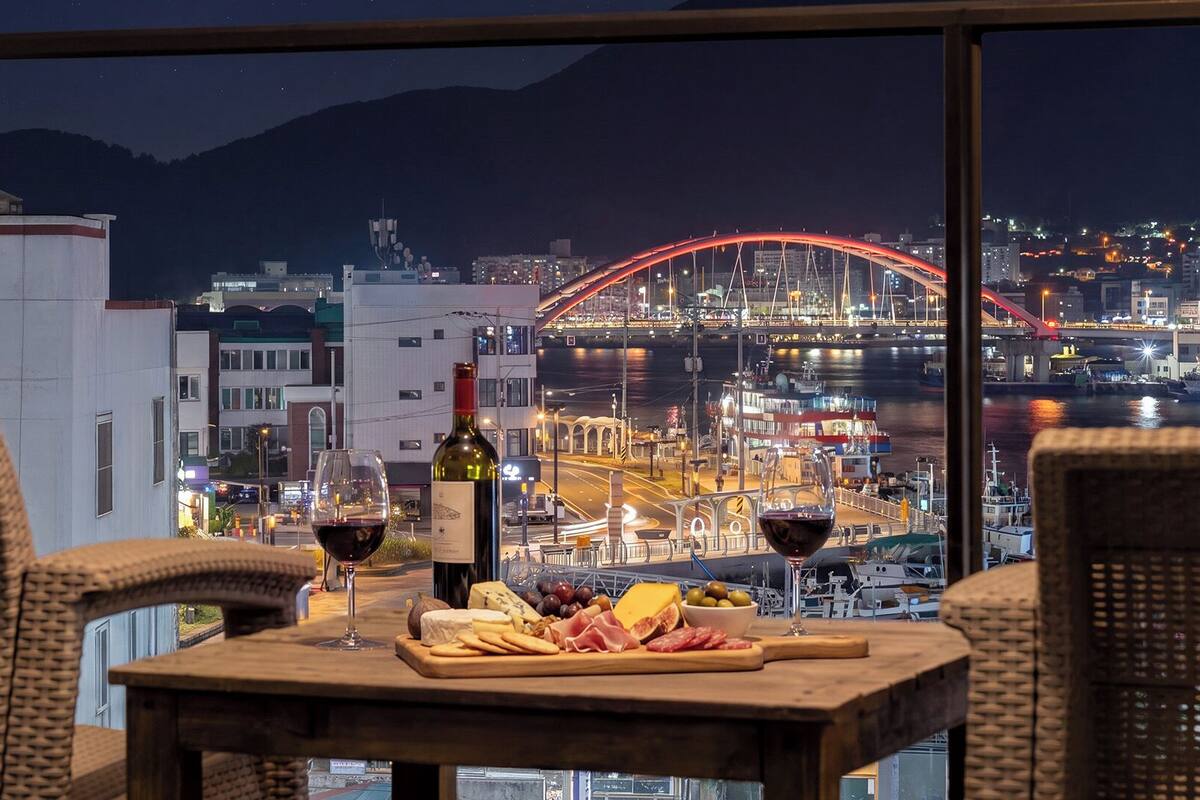Una mesa con una botella de vino, copas, queso y embutidos, con vistas a un paisaje urbano que incluye un puente y edificios.