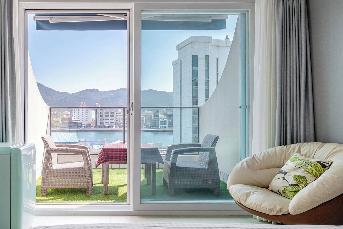 Un balcón con vistas al agua y edificios, amueblado con sillas de mimbre y una mesa.