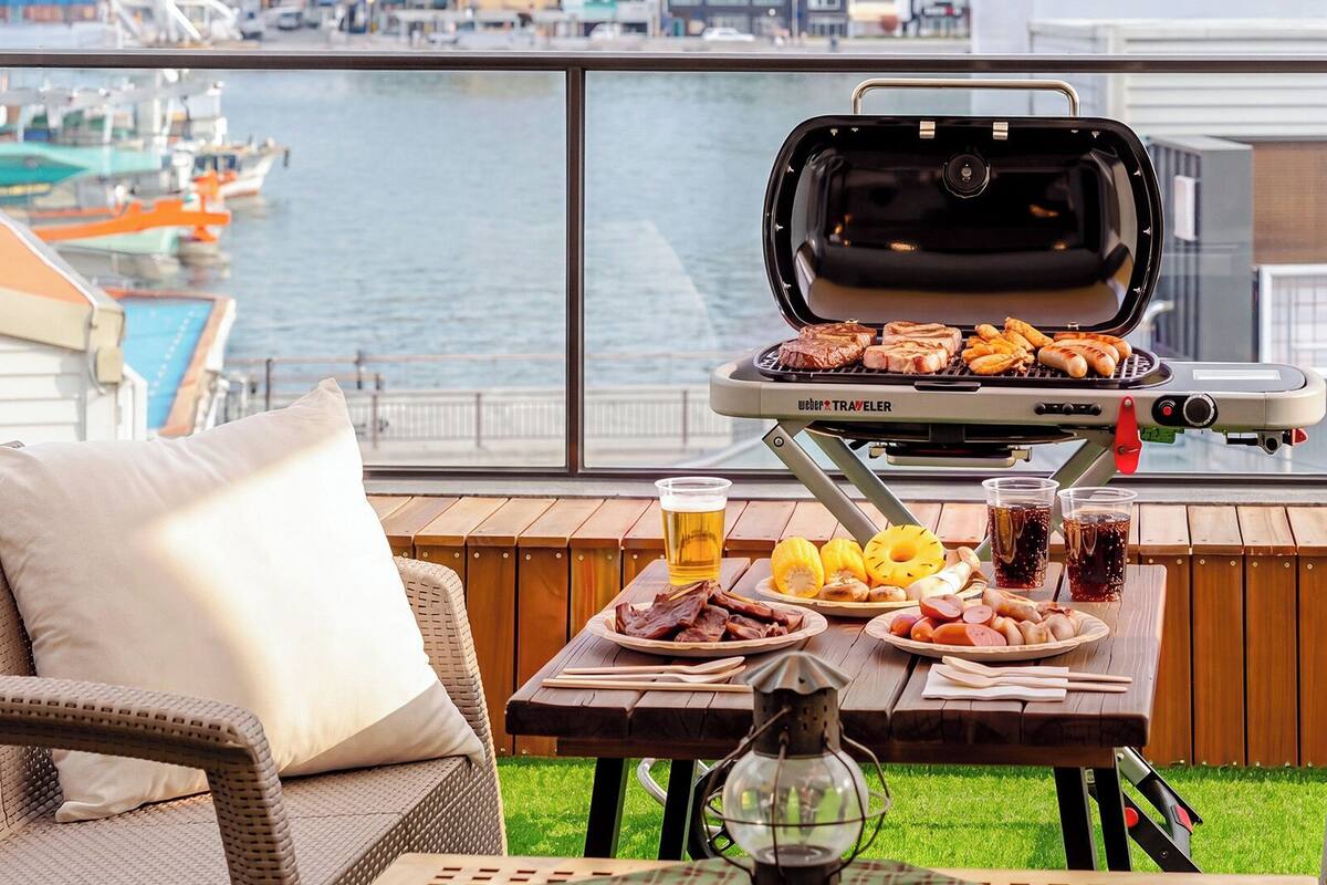 Un balcón con una parrilla, una silla de mimbre, una mesa con comida y bebidas, y vista al puerto deportivo.