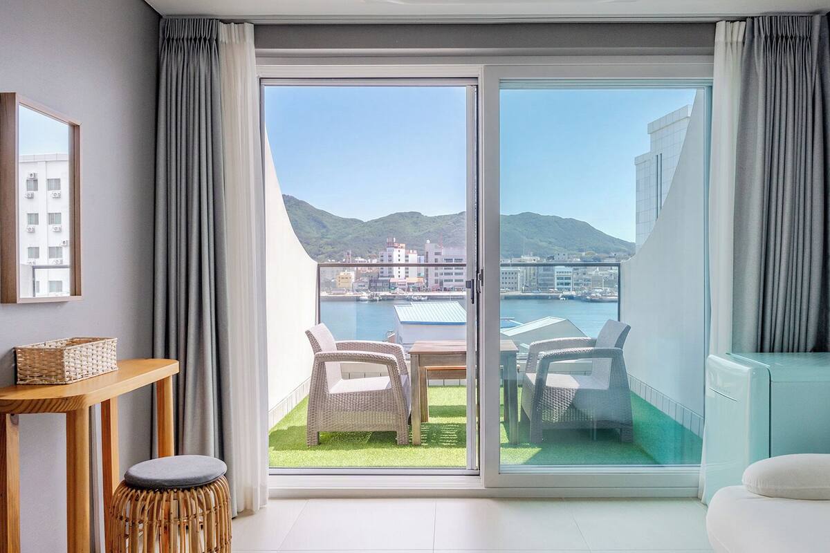 Una habitación con puerta corrediza de vidrio que da a un balcón con mobiliario exterior y vistas a un paisaje urbano y colinas.
