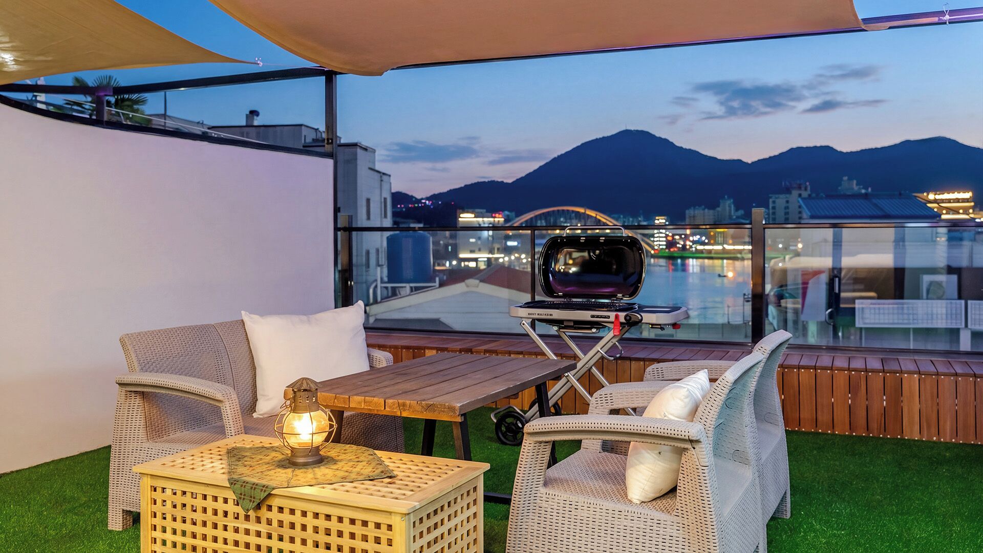 A rooftop terrace with outdoor furniture, a grill, and a view of a cityscape and mountains.