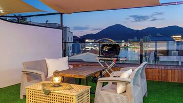 A rooftop terrace with outdoor furniture, a grill, and a view of a cityscape and mountains.