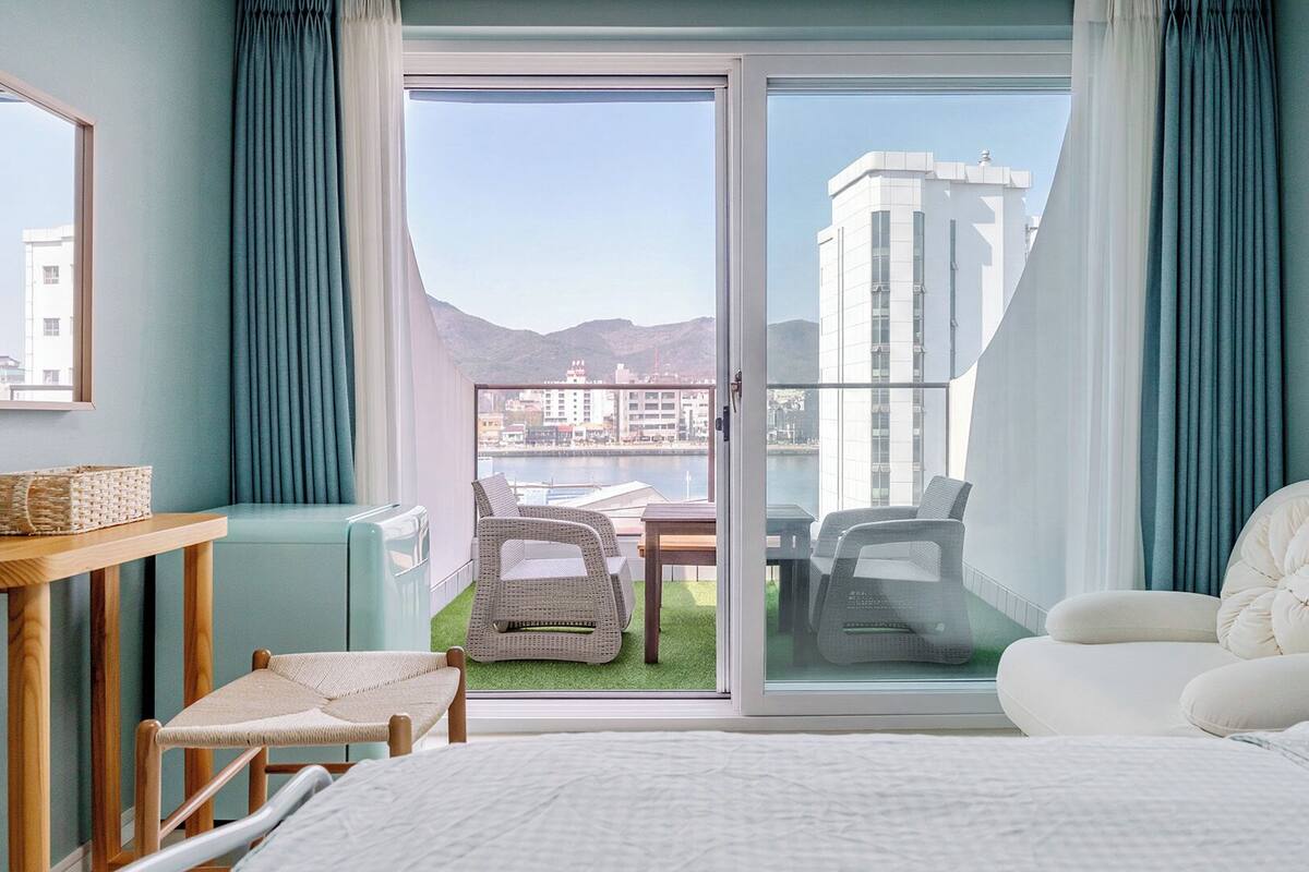 Habitación de hotel con cama, zona de estar con sillas, una mesa y vistas a la ciudad y montañas a través de un amplio ventanal corredera.