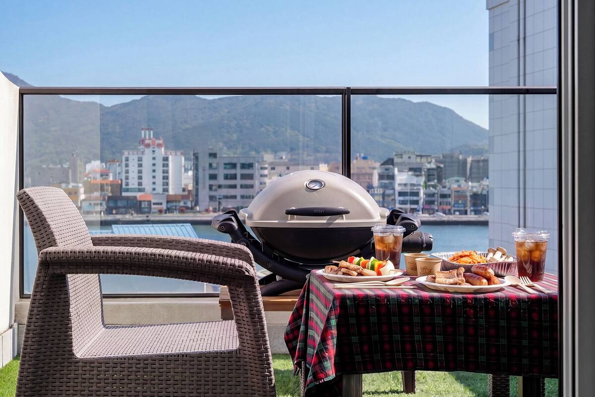 Un balcón con una parrilla, una silla de mimbre, una mesa con comida y vistas a la ciudad.