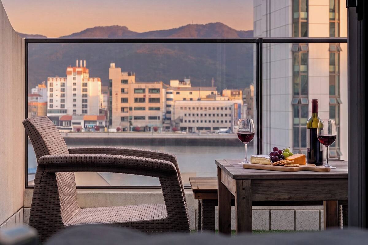 Un balcón con una silla de mimbre, una mesita con una botella de vino, una copa de vino, queso, uvas y otra botella de vino.