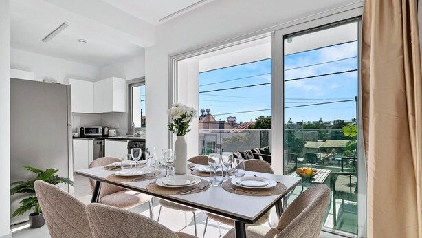 Ein moderner Essbereich mit einem großen Tisch, gedeckt für vier Personen, einer Küche im Hintergrund und Blick auf die Stadt durch große Fenster.