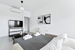 A modern hotel room with a flat-screen TV, a bed with a grey quilt, a white chair, and abstract wall art.