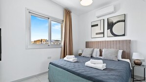 Ein modernes Schlafzimmer mit einem großen Bett, einem Fenster mit Vorhängen, einer Wand-Klimaanlage und zwei gerahmten Kunstwerken.