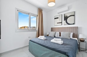 A modern bedroom with a large bed, a window with curtains, a wall-mounted air conditioner, and two framed artworks.