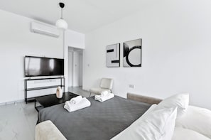 A modern hotel room with a flat-screen TV, a bed with a grey quilt, a white chair, and abstract wall art.