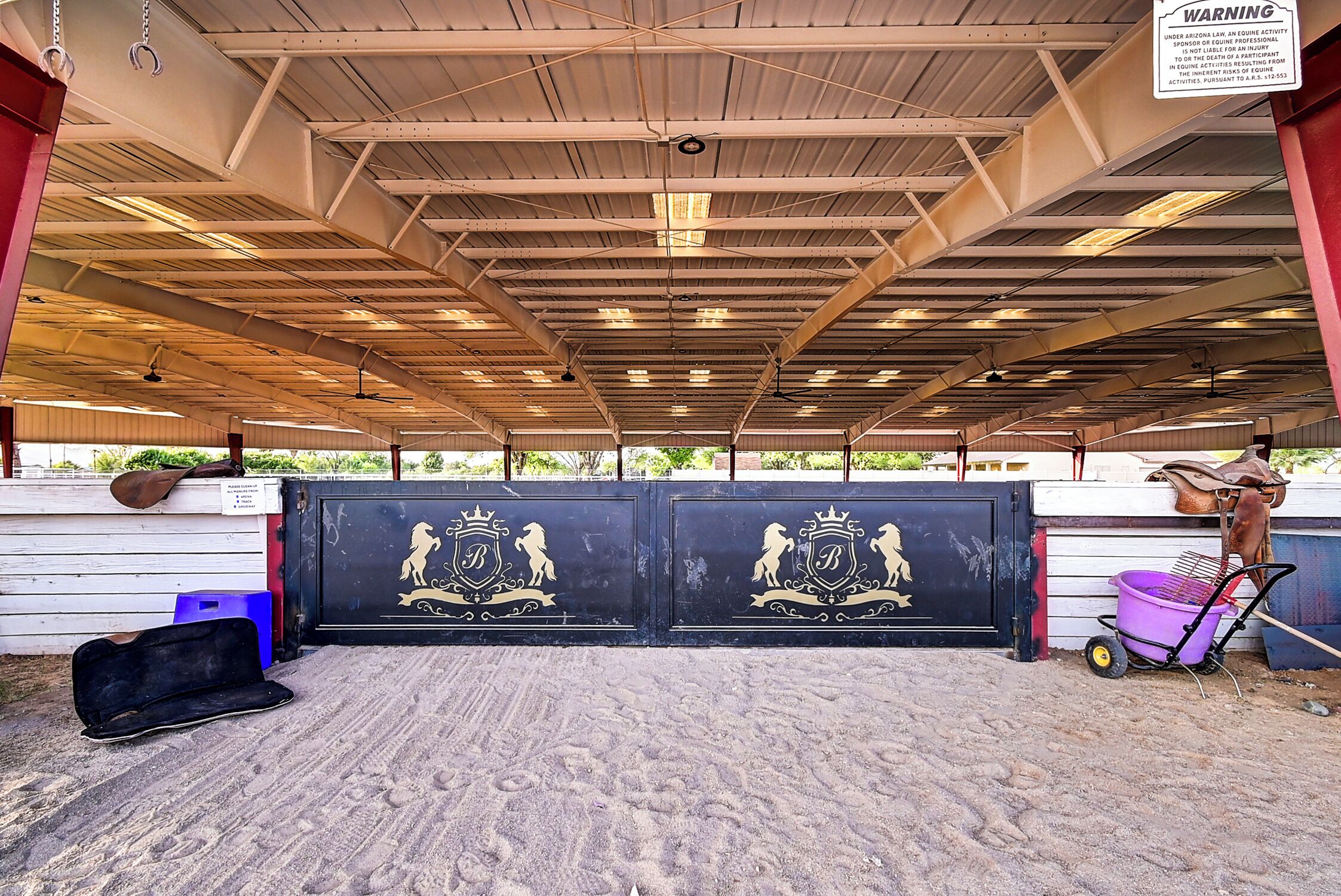 An indoor riding arena with a metal roof, a black gate with a crest, and a wheelbarrow.