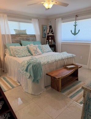 A bedroom with a bed, a wooden side table, a shelf with decorative items, and a view of the sea through a window. - Isles of Carpri Charming 2 bedroom  home with heated pool and boat dock. (Naples)
