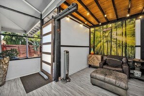 A room with a sliding glass door, a standing fan, a leather sofa, and a large forest mural. - Serene 2BR Valley Glen Oasis: Private Yard, & BBQ (Los Angeles)
