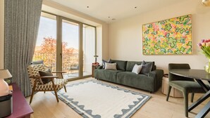A living room with a sofa, armchair, and dining table, large windows with a view, and a colorful abstract painting on the wall. - Balham Modern - Stylish 1bed Flat With Balcony (Moffat)