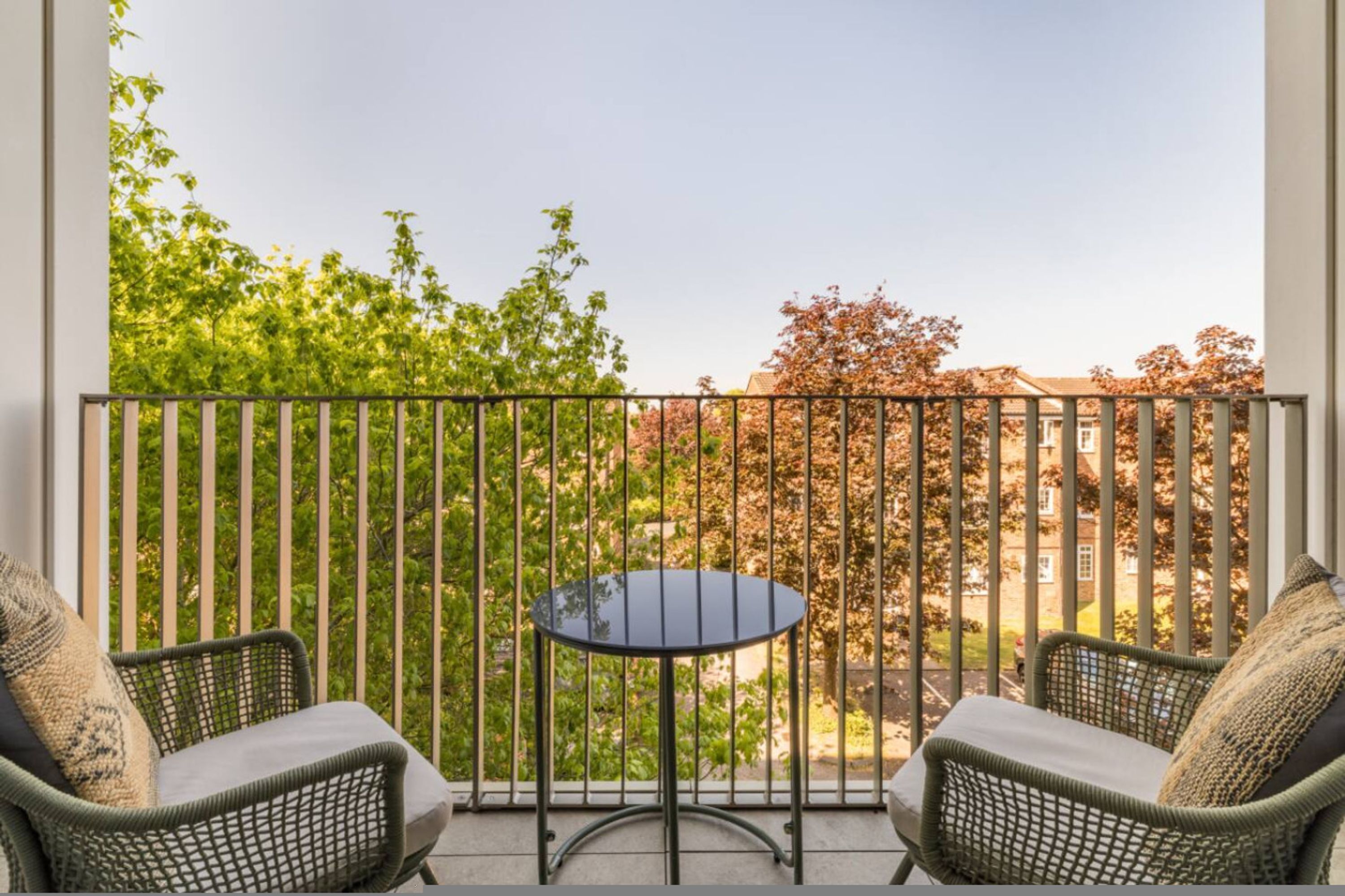 A balcony with two wicker chairs and a small round table, overlooking a residential area with trees and houses.
