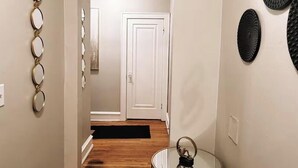 Un pasillo con suelo de madera, una puerta blanca y una mesa de vidrio con un objeto negro encima.