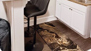 Cocina moderna con armarios blancos, una silla negra, alfombra con estampado de mármol y un adorno en la pared.