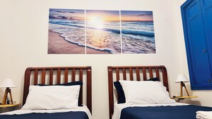 Une chambre d’hôtel avec deux lits, une porte bleue et un tableau représentant un coucher de soleil accroché au mur.