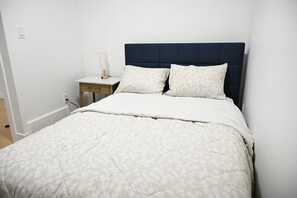 A neatly made bed with a floral-patterned bedspread and two pillows, a bedside table with a lamp, and a wall socket. - Relax in this calm, spacious, stylish space featuring top-of-the-line appliances (Nisku)